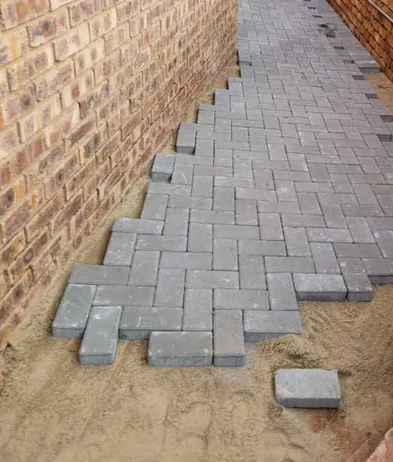 Types of Paving Bricks in South Africa 2 Plain rectangular grey pavers lying loose on bedding sand at the stepped leading edge of a herringbone field, one unit on its own with its outline and chamfered top edge visible